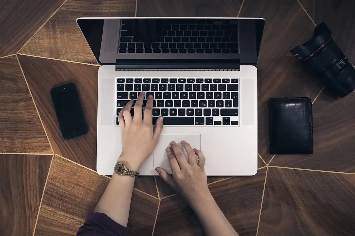 Mâini pe laptop, văzute de sus, cu un telefon și o cameră alături
