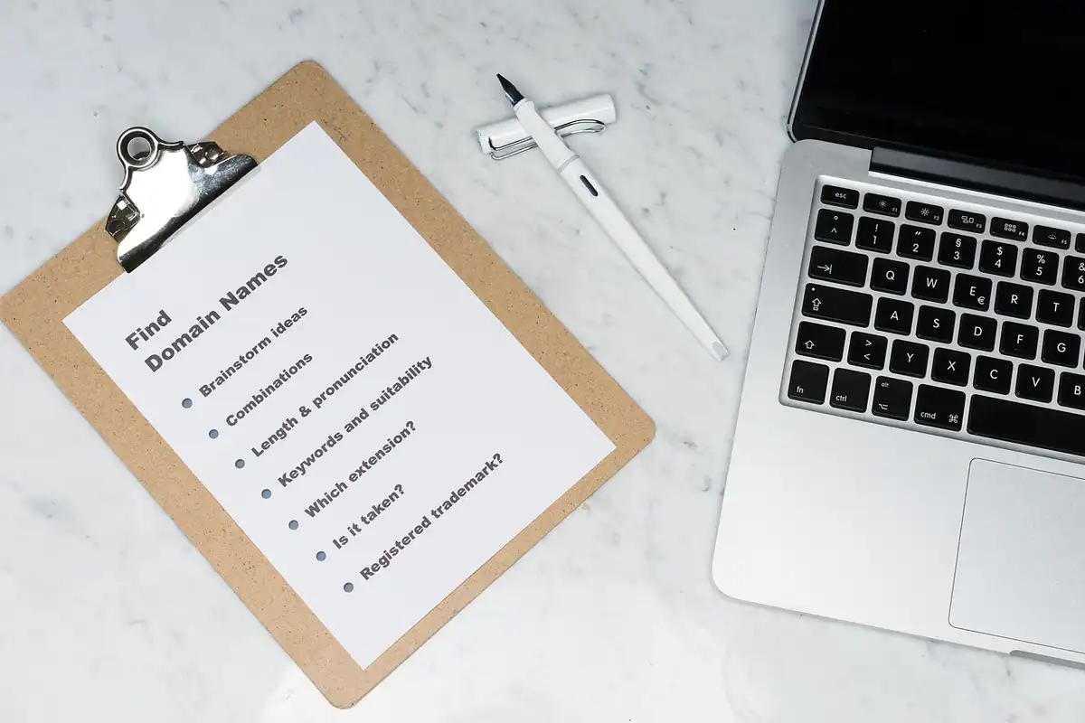 Clipboard cu o listă de idei pentru numele de domeniu, lângă un laptop
