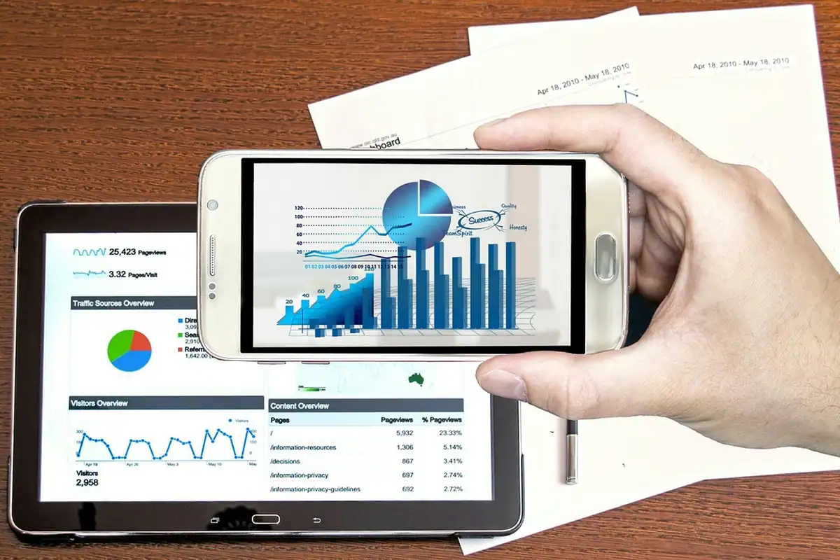 Mână care ține un telefon cu grafice, lângă o tabletă cu statistici