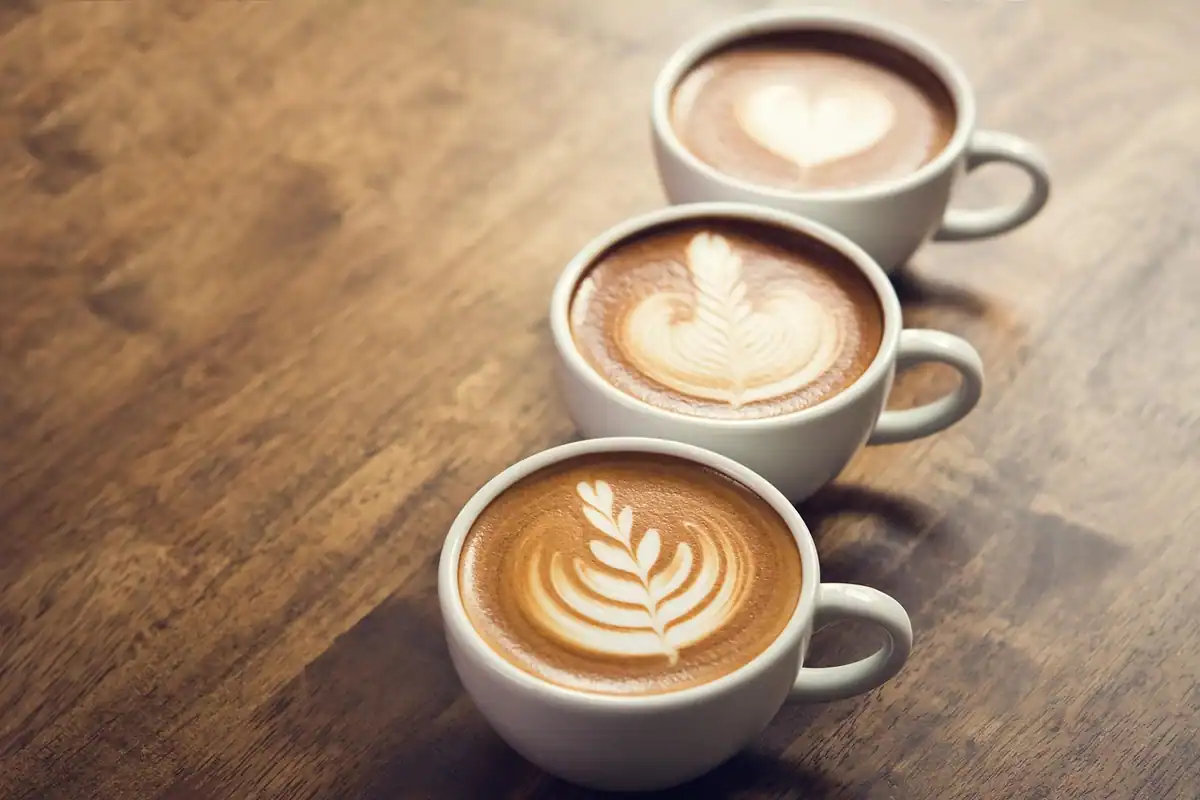 Trei cești de cafea cu desene în spumă, așezate pe o masă de lemn