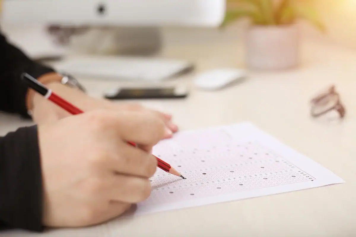 Mână care completează un formular cu creionul, pe o masă de lemn