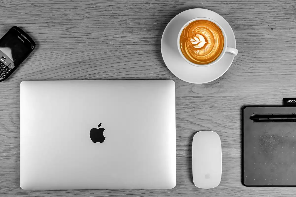 Laptop argintiu, cafea și mouse pe un birou, într-o fotografie alb-negru