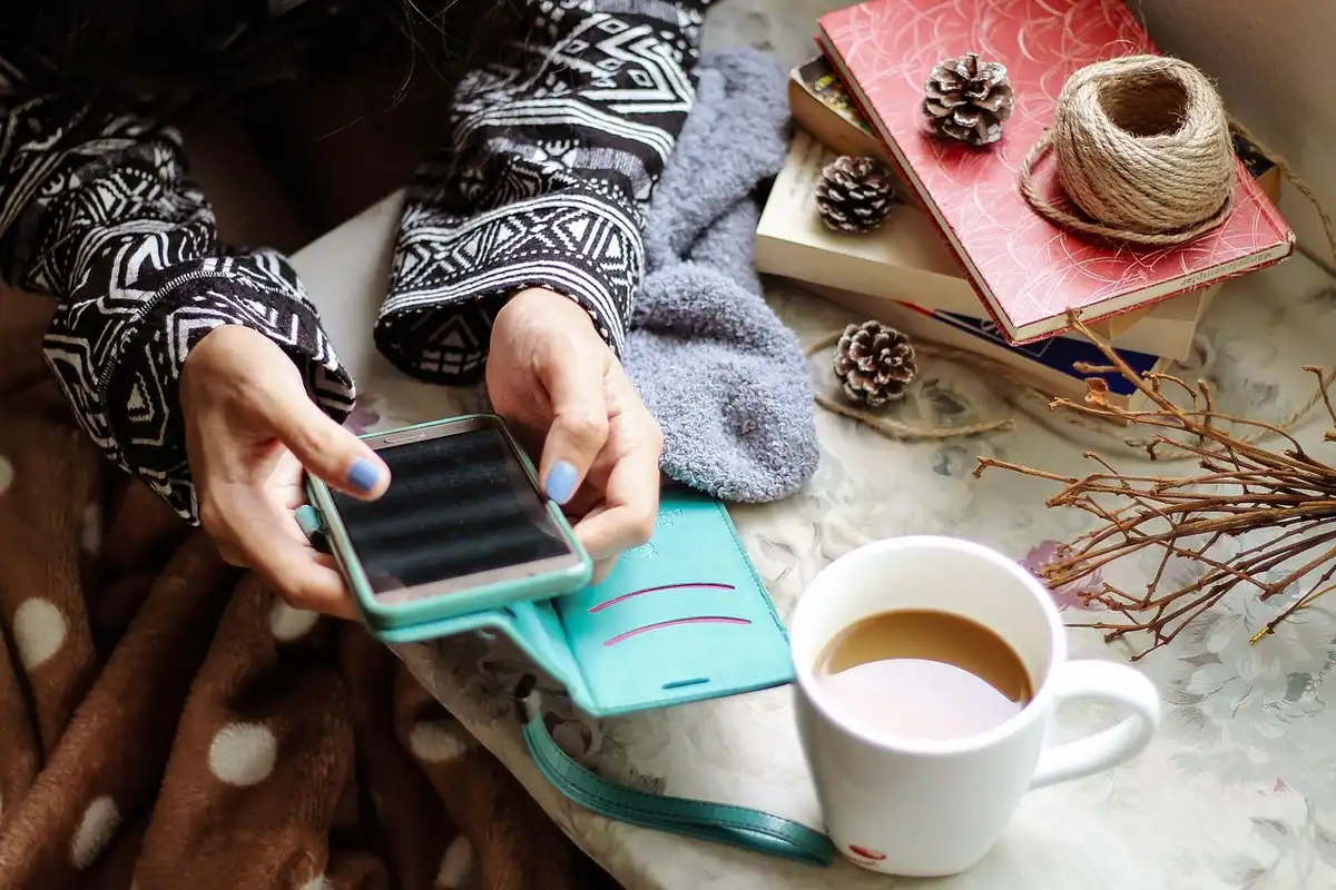Mâini care țin un telefon, lângă o cană cu cafea și cărți