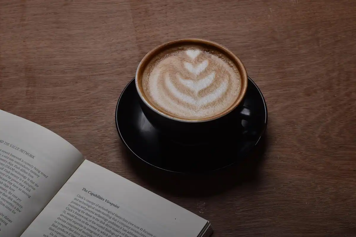 Cappuccino cu spumă lângă o carte deschisă, pe o masă din lemn
