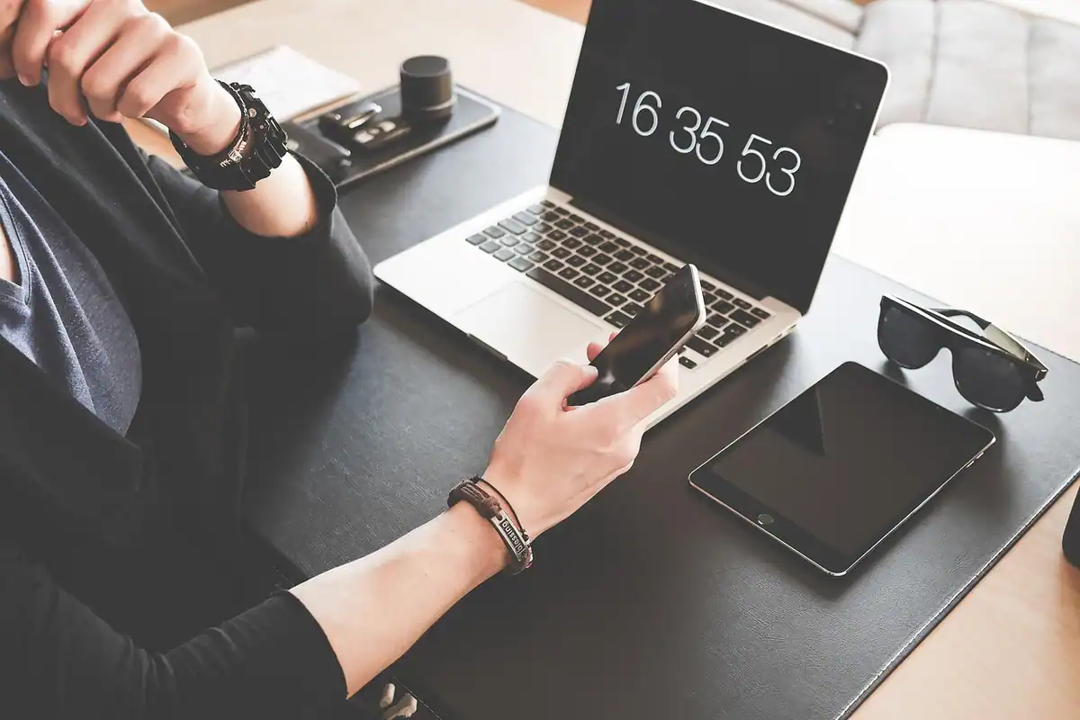 Mâini care țin un telefon, lângă un laptop, o tabletă și ochelari de soare
