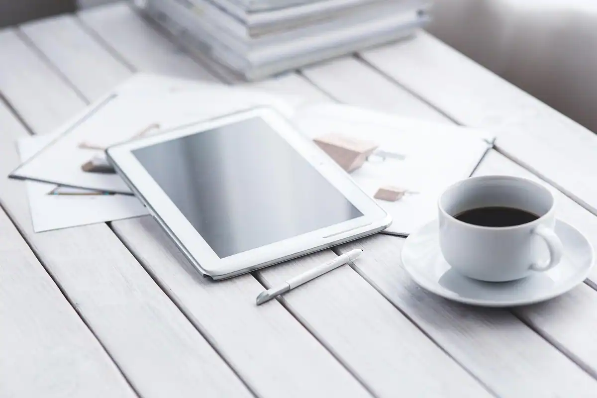 Tabletă albă, cafea și foi așezate pe o masă albă din lemn
